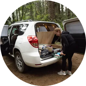 A woman is getting something out of the back of a car from the storage compartment of a camper set up.