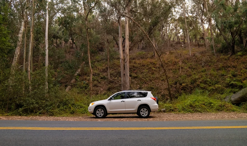 A side shot of a white Rav4 on the side of the road in front of a hill and trees.